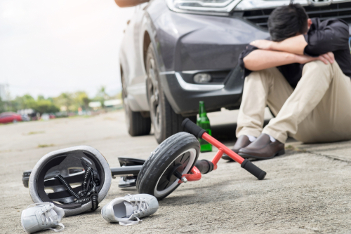 Distraught man sitting on the ground next to a damaged car and child's bicycle with helmet and shoes scattered around.