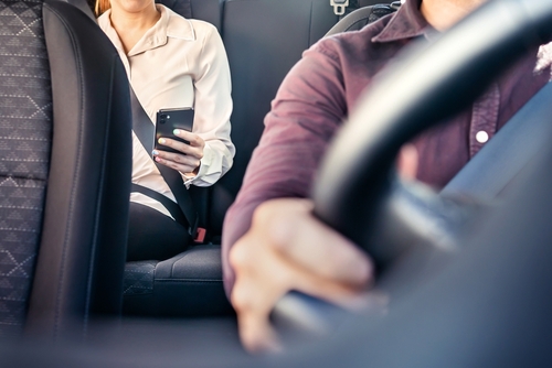 View inside a car showing a male driver steering and a female passenger using a smartphone, depicting a typical travel scene.