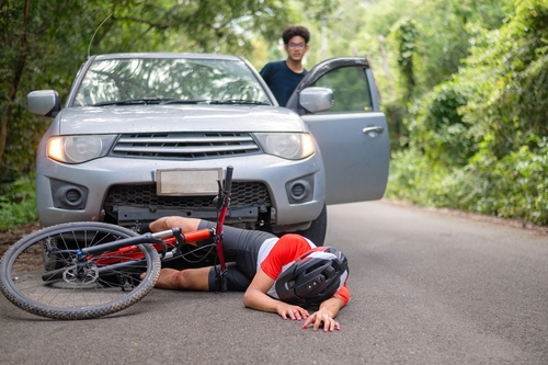 A bicyclist lying on the road next to his overturned bike, injured after being hit by a car, with the car driver standing by the open door, looking concerned