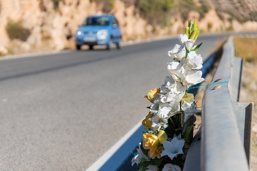 Floral tribute attached to a guardrail along a scenic road, with a blue car passing by in the background.