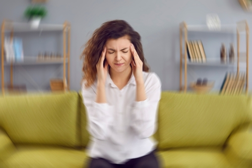 A young woman experiencing a severe headache, sitting on a yellow sofa in a modern living room.