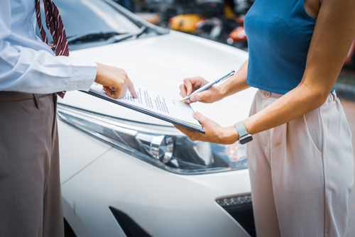 Close-up of a female client signing insurance or repair documents on a clipboard, presented by a male insurance agent beside a car.