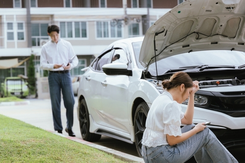 A woman sitting by a broken-down car with the hood open, looking distressed, while a man stands beside her taking notes.