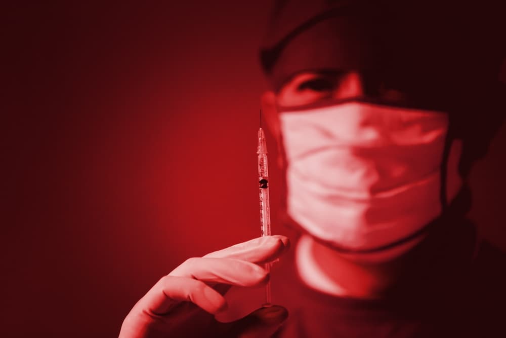 A masked medical professional holding a syringe, with the image filtered in a red tone.