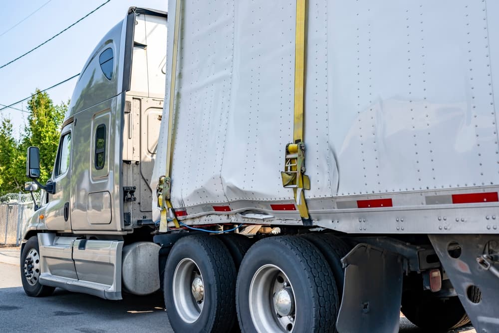 A damaged semi-truck trailer with visible dents on the side, secured with yellow straps.