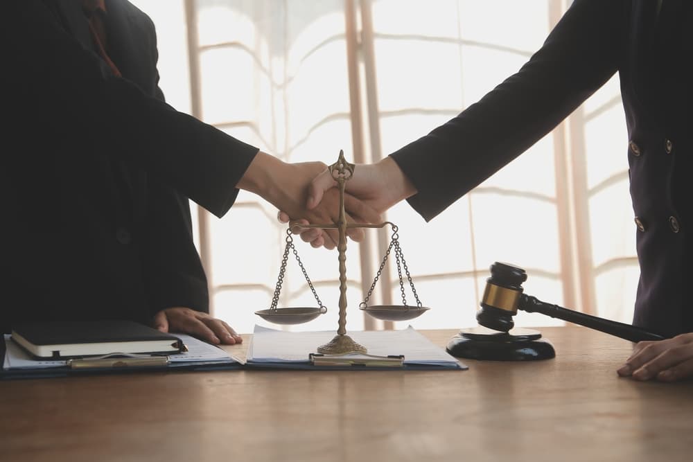 Two individuals shaking hands over a legal desk with scales of justice and a gavel.