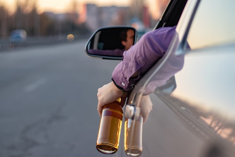 A driver extending their arm out of a car window, holding a bottle of alcohol.