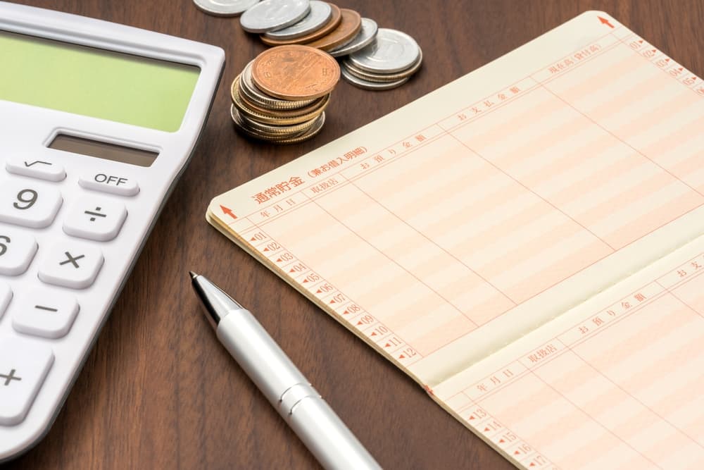 A calculator, coins, a bank book, and a pen placed on a wooden table surface.